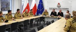 Szersze ujęcie narady w Komendzie Wojewódzkiej PSP w Katowicach. Przy dużym stole siedzi kilkunastu funkcjonariuszy w piaskowych koszulach służbowych oraz oficerowie w mundurach galowych. W tle znajdują się flagi Polski i Unii Europejskiej oraz tablica z logo Państwowej Straży Pożarnej. Spotkanie odbywa się w jasnej, nowoczesnej sali konferencyjnej.