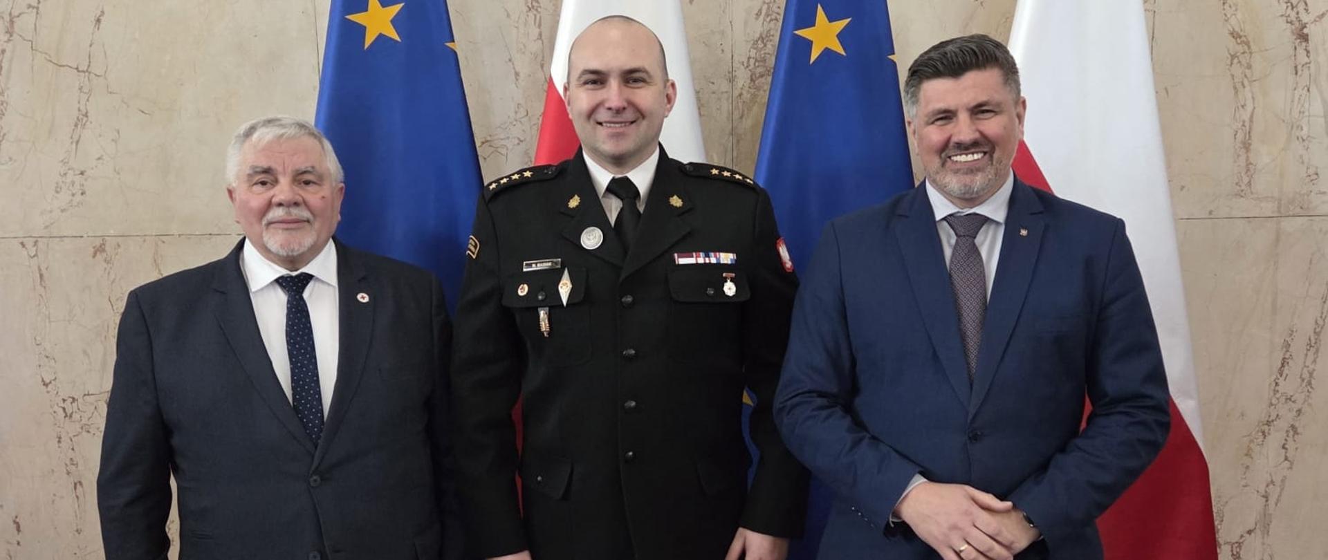 Trzej mężczyźni na czerwonym dywanie, ustawieni w jednym rzędzie i zwróceni przodem do aparatu. W tle widoczne są ustawione naprzemiennie cztery flagi - dwie Polski (biało-czerwone) oraz dwie Unii Europejskiej (niebieskie z żółtymi gwiazdami). Mężczyzna po lewej to starszy pan w ciemnym garniturze, białej koszuli i krawacie, z siwymi włosami i wąsami. W środku stoi mężczyzna w galowym mundurze strażackim z odznaczeniami i dystynkcjami, uśmiechnięty. Po prawej znajduje się mężczyzna w granatowym garniturze, białej koszuli i krawacie, również uśmiechnięty