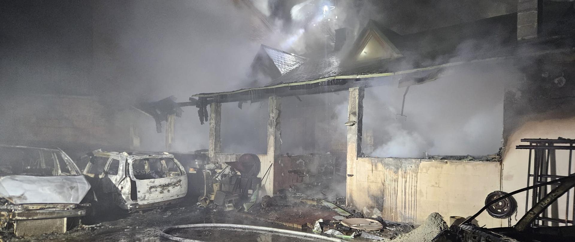 Na zdjęciu widać pogorzelisko po nocnym pożarze: spalony budynek z zawalonym zadaszeniem, unoszący się gęsty dym oraz zwęglone wraki kilku samochodów stojących na mokrej, osmolonej ziemi. Przed obiektem leżą rozciągnięte węże gaśnicze, a cała scena jest oświetlona silnym światłem ratowniczym przebijającym się przez dym.