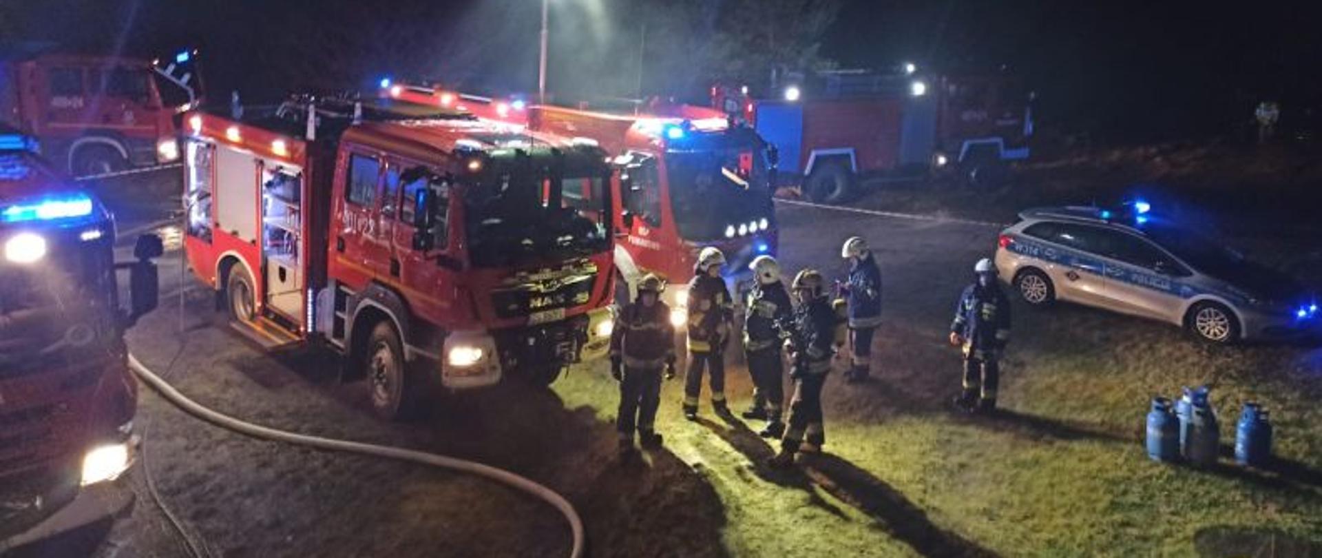 Zdjęcie przedstawia miejsce koncentracji sił biorących w działaniu gaśniczym. Zdjęcie wykonane z podwyższonej pozycji. Centralny punkt stanowią strażacy i pojazdy gaśnicze. Zdjęcie oświetlone najasnicami strażackimi. Na zdjęciu również radiowóz policyjny.