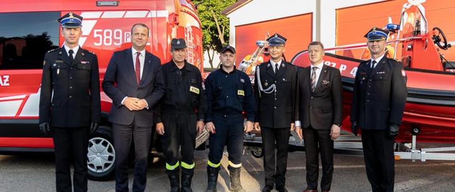 grupa strażaków i mnężczyzn w ubraniach cywilnych stoi w szeregu, zdjęcie grupowe, w tle czerwony samochód strażacki oraz łódź