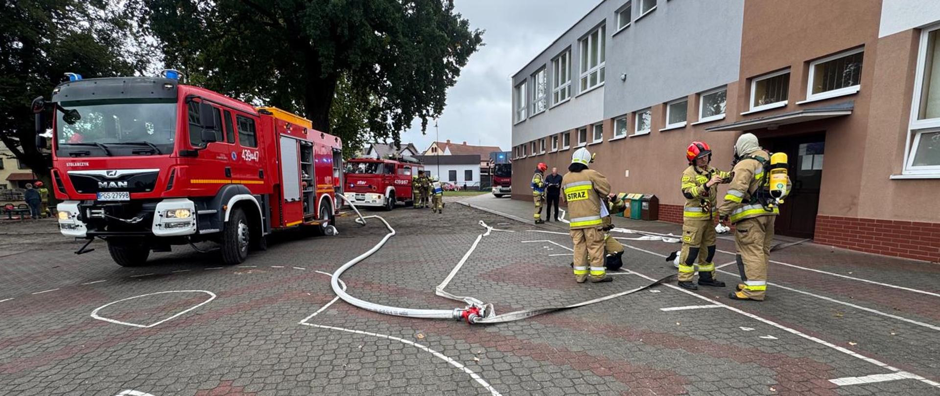 Trzy wozy strażackie na placu wewnętrznym przed budynkiem szkoły. Z wozów rozwinięta jest linia gaśnicza. Przed wejściem stoją strażacy.