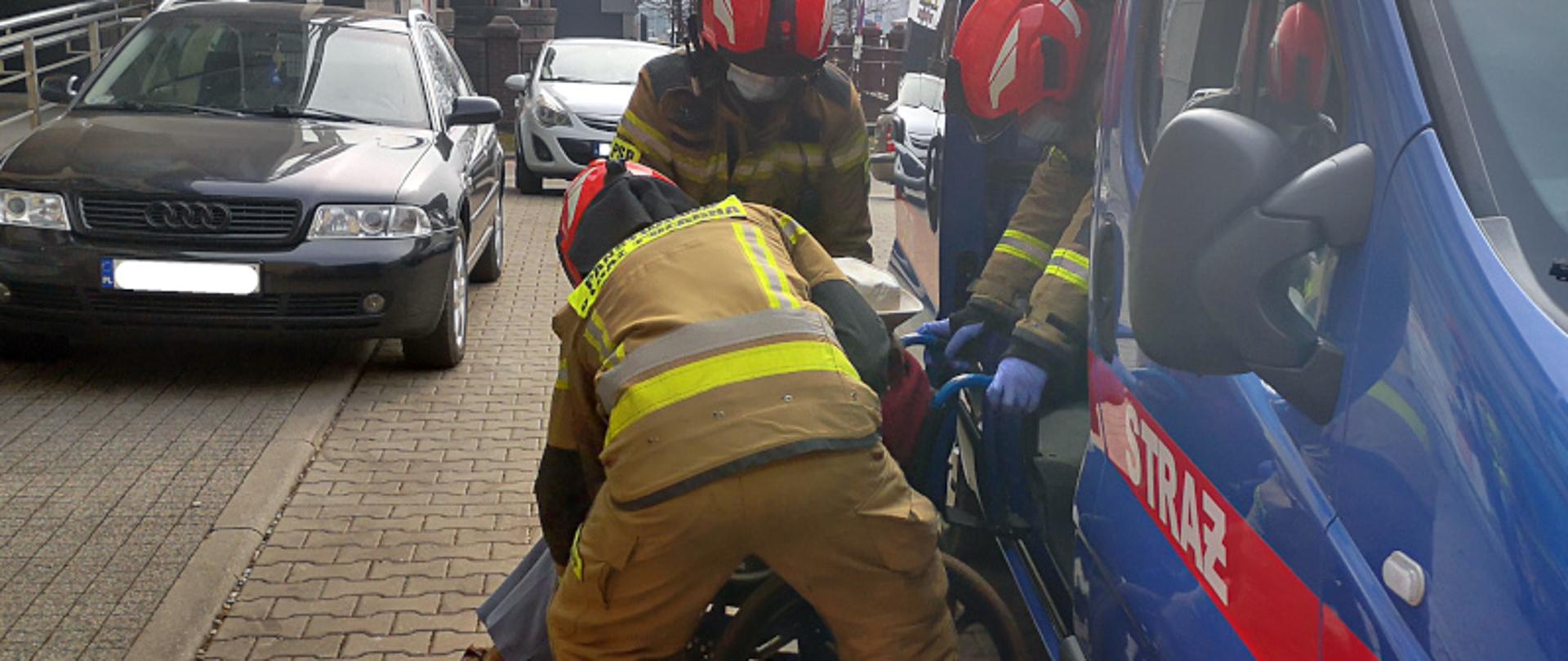 Strażacy pomagający osobie poruszającej się na wózku inwalidzkim wysiąść z samochodu, pod budynkiem poradni lekarskiej