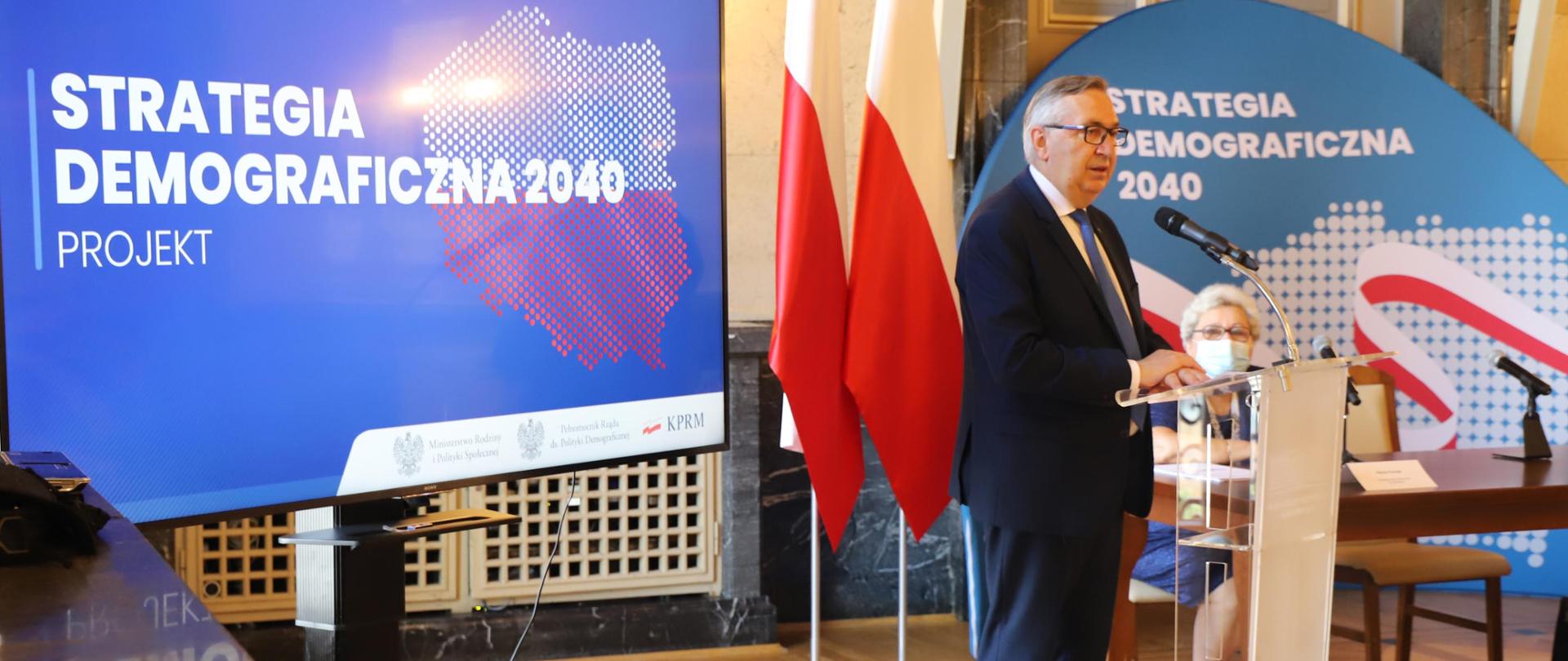 Minister S. Szwed stojący przy mównicy. W tle stojące flagi narodowe oraz baner z napisem Strategia Demograficzna 2040. Projekt. W tle kontury Polski.