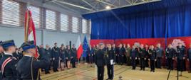 Uroczystość w hali sportowej z udziałem przedstawicieli służb mundurowych i cywilnych. Na pierwszym planie dwóch oficerów Państwowej Straży Pożarnej stoi naprzeciw siebie — jeden z nich trzyma dokumenty, drugi znajduje się w pozycji na baczność. Po lewej stronie widoczni są funkcjonariusze w czarnych mundurach salutujący do sztandaru z orłem białym i napisem „Bogu na chwałę, ludziom na ratunek”. W tle znajdują się flagi Polski i Unii Europejskiej oraz szeregi zaproszonych gości w mundurach i garniturach, stojących na tle czerwonych tablic z emblematem PSP.