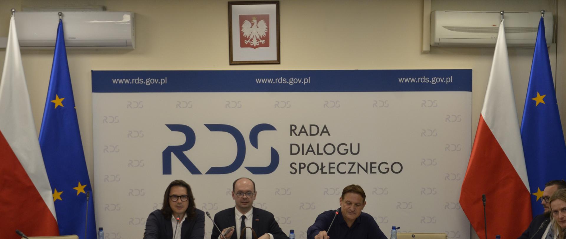 Zdjęcie przedstawia posiedzenie połączonych zespołów problemowych ds. międzynarodowych i ds. prawa pracy. Na pierwszym planie widoczny jest stół prezydialny, za którym siedzą przewodniczący zespołu Błażej Mądrzycki oraz wiceminister Sebastian Gajewski oraz przedstawiciel Konfederacji Lewiatan Jacek Męcina. Za nimi stoi banner z napisem Rada Dialogu Społecznego, a po jego obu stronach stoją flagi Polski i Unii Europejskiej. 