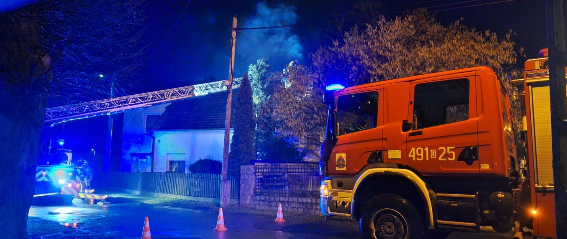 Na zdjęciu znajdują się samochody strażackie, w tym drabina, widać dym unoszący się z komina w budynku mieszkalnym