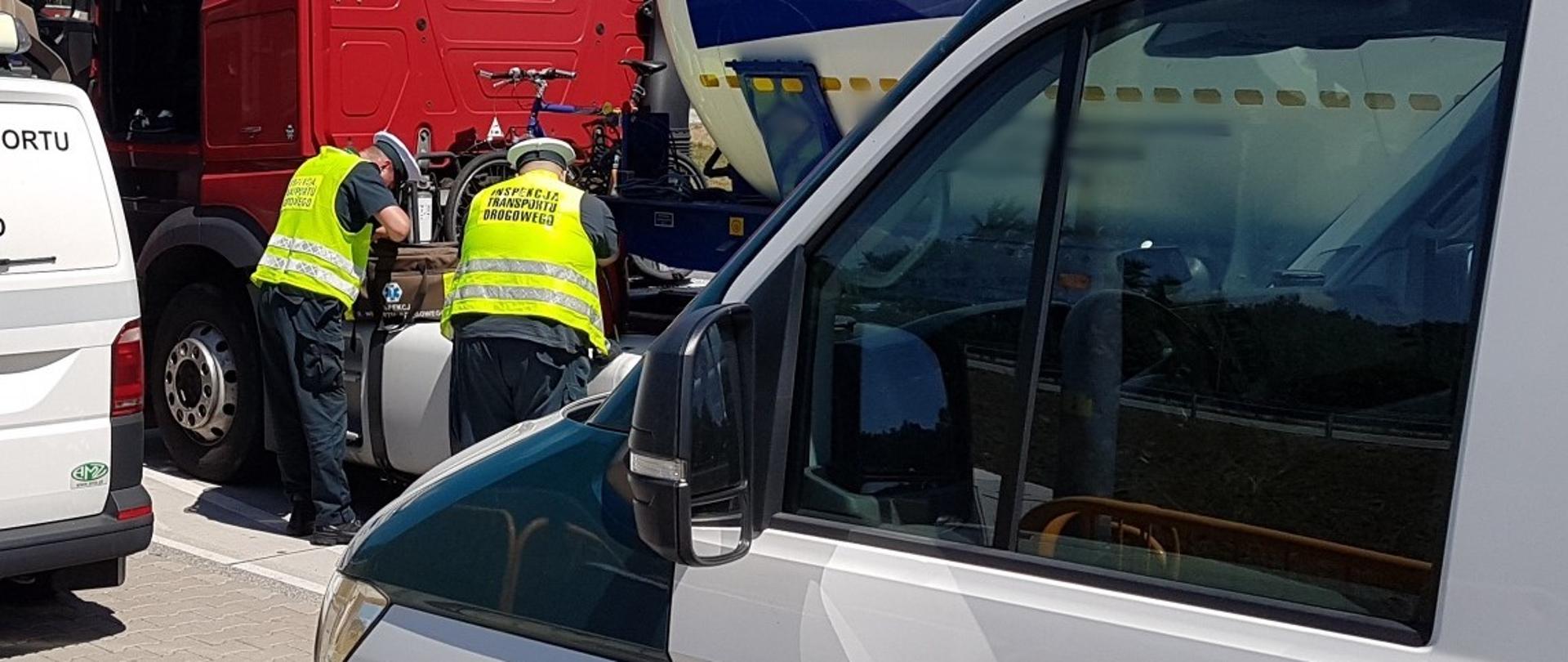 Na pierwszym planie przód oznakowanego radiowozu ITD typu furgon. W tle dwóch inspektorów ITD stoi obok ciężarówki i wyjmuje środki do udzielenia pierwszej pomocy z torby medycznej.
