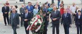 Delegacja kobiet i mężczyzn składa wieniec. Na pierwszym planie mężczyzna w mundurze niesie wieniec. 