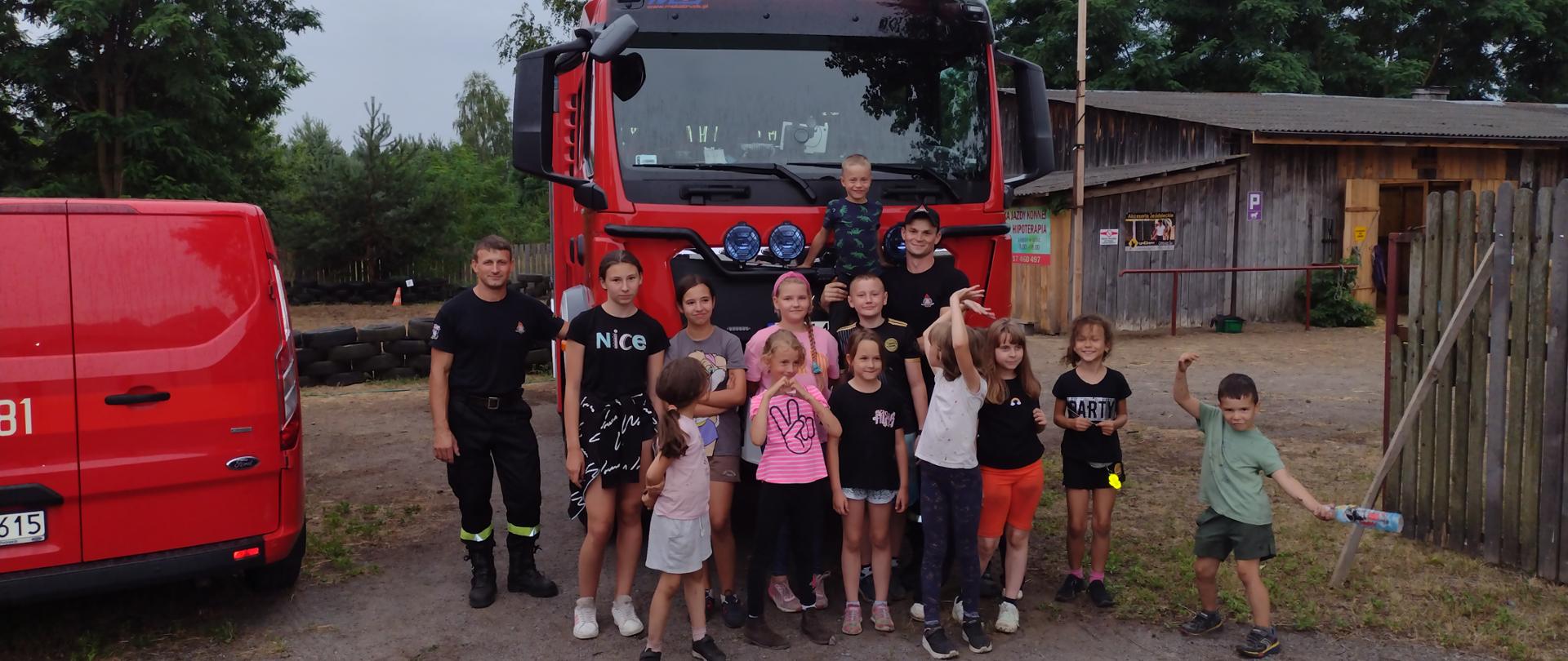 Zdjęcie grupowe dzieci wraz ze strażakami na tle samochodu gaśniczego i budynków stadniny.