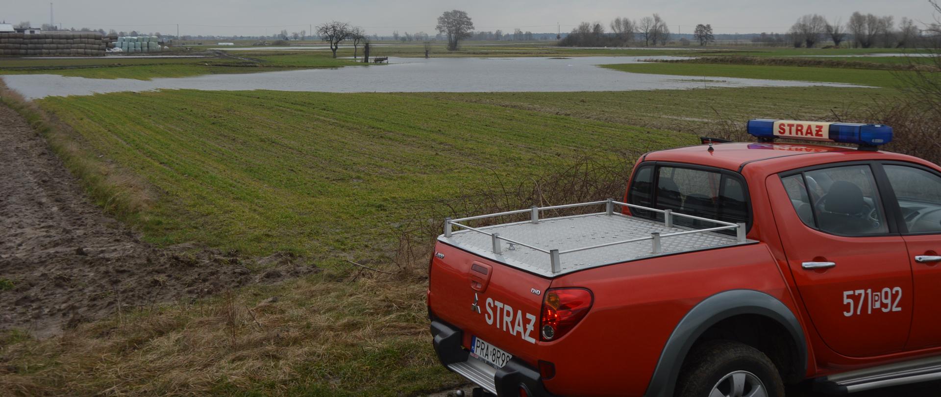 Okolice miejscowości Wydawy. Na pierwszym planie samochód terenowy straży pożarnej. Dalej grunty rolne, cześciowo znajdujące się pod wodą. W tle drzewa, sterty słomy oraz linia energetyczna.