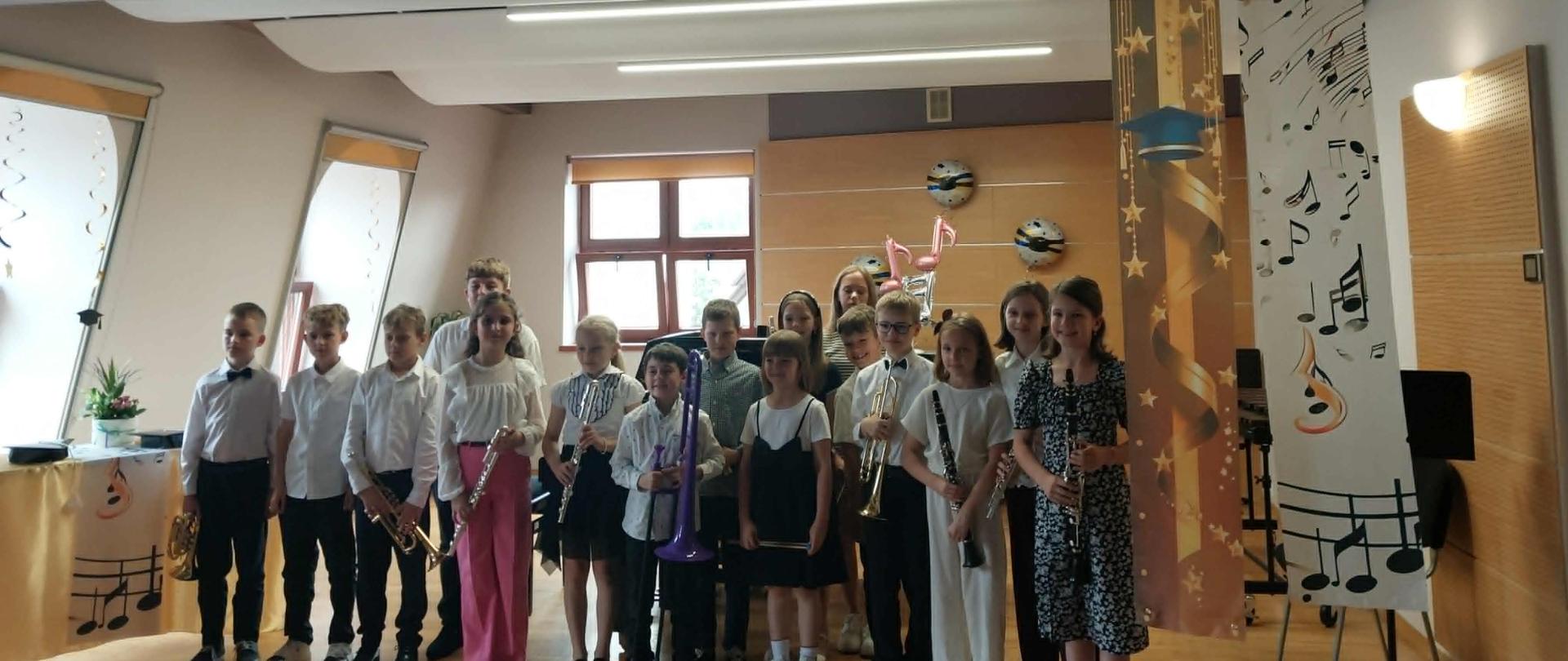 Zdjęcie przedstawia grupę kilkunastu dzieci stojących na scenie w sali lub szkolnej, pozujących do wspólnej fotografii.
Uczestnicy: Dzieci są elegancko ubrane – chłopcy w białe koszule (niektórzy z muszkami), a dziewczynki w odświętne bluzki, sukienki lub spódnice. Część z nich trzyma w dłoniach instrumenty muzyczne, takie jak flety, trąbka czy klarnet.
Otoczenie: W tle widoczne są elementy dekoracyjne związane z uroczystością lub motywami muzycznymi – wiszące banery (jeden z motywem czapki absolwenta, drugi z nutami) oraz balony w kształcie nut. Po lewej stronie widać fragment stołu z dekoracyjnym obrusem. Scena ma drewnianą podłogę, a pomieszczenie jest nowocześnie oświetlone podłużnymi lampami sufitowymi.
