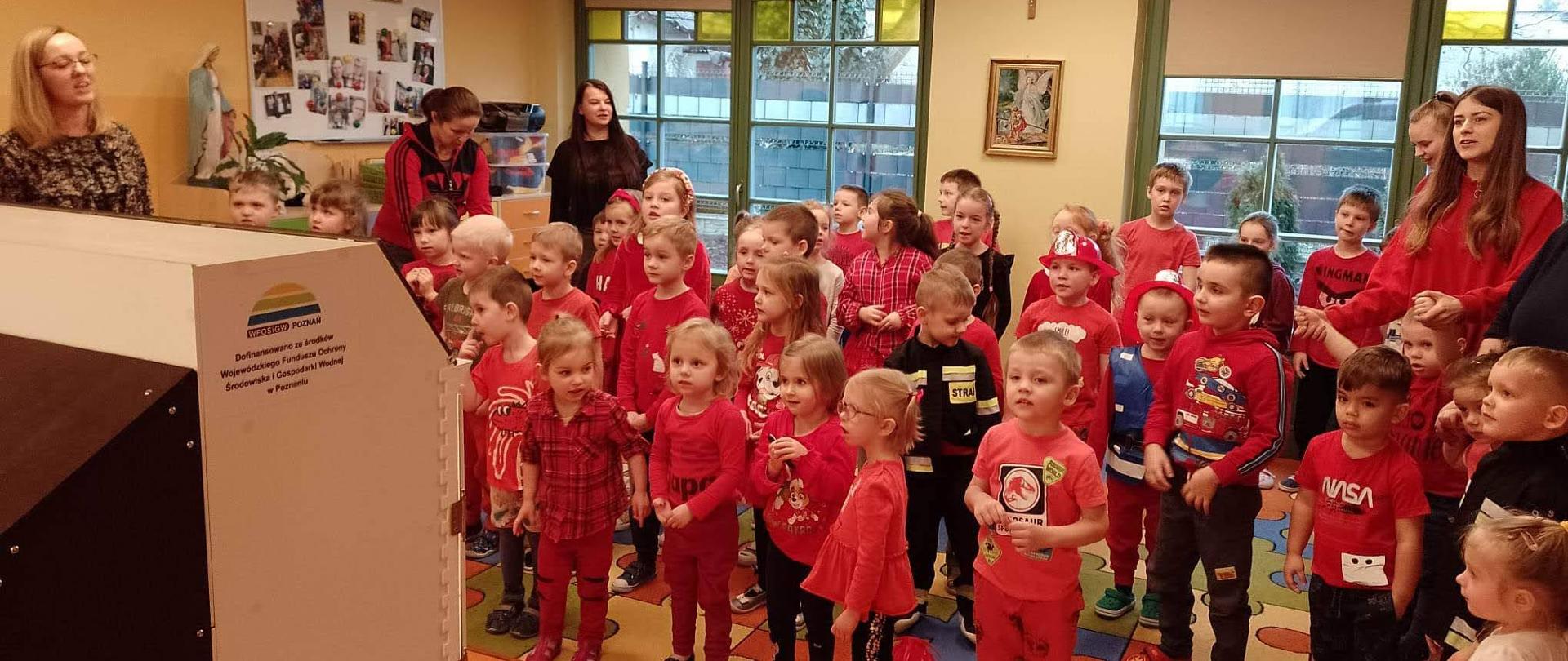 Na zdjęciu grupa dzieci w wieku przedszkolnym w sali lekcyjnej. Dzieci ubrane na czerwono czekają na prelekcje strażaków z użyciem symulatora zagrożeń. W sali znajdują się również opiekunowie najmłodszych