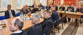 Zdjęcie przedstawia słuchaczy narady zajmujących miejsce przy stole, w tle widać gablotę z pucharami oraz banery państwowej straży pożarnej koloru czerwonego.