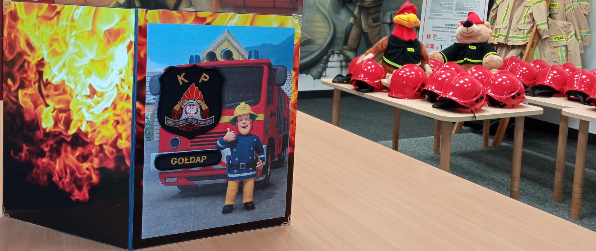 Zdjęcie przedstawia stronę frontową oraz tył kartki z okazji Dnia Dziecka z życzeniami od gołdapskich strażaków zlokalizowanej w sali edukacyjnej dla dzieci Komendy Powiatowej Państwowej Straży Pożarnej w Gołdapi