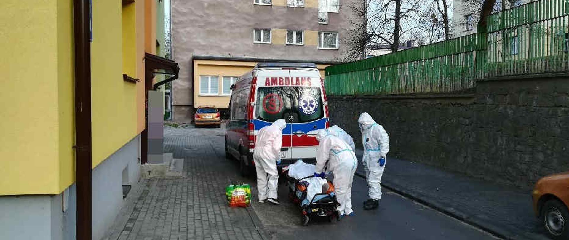 Zdjęcie zrobione na parkingu między blokami. Na zdjęciu widać karetkę pogotowania ratunkowego. Obok karetki widać czterech ratowników ubranych w białe barania ochronne przed COVID-19. Ratownicy transportują osobę chorą do karetki. W oddali widać samochód osobowy, który stoi obok bloku. 