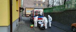 Zdjęcie zrobione na parkingu między blokami. Na zdjęciu widać karetkę pogotowania ratunkowego. Obok karetki widać czterech ratowników ubranych w białe barania ochronne przed COVID-19. Ratownicy transportują osobę chorą do karetki. W oddali widać samochód osobowy, który stoi obok bloku. 