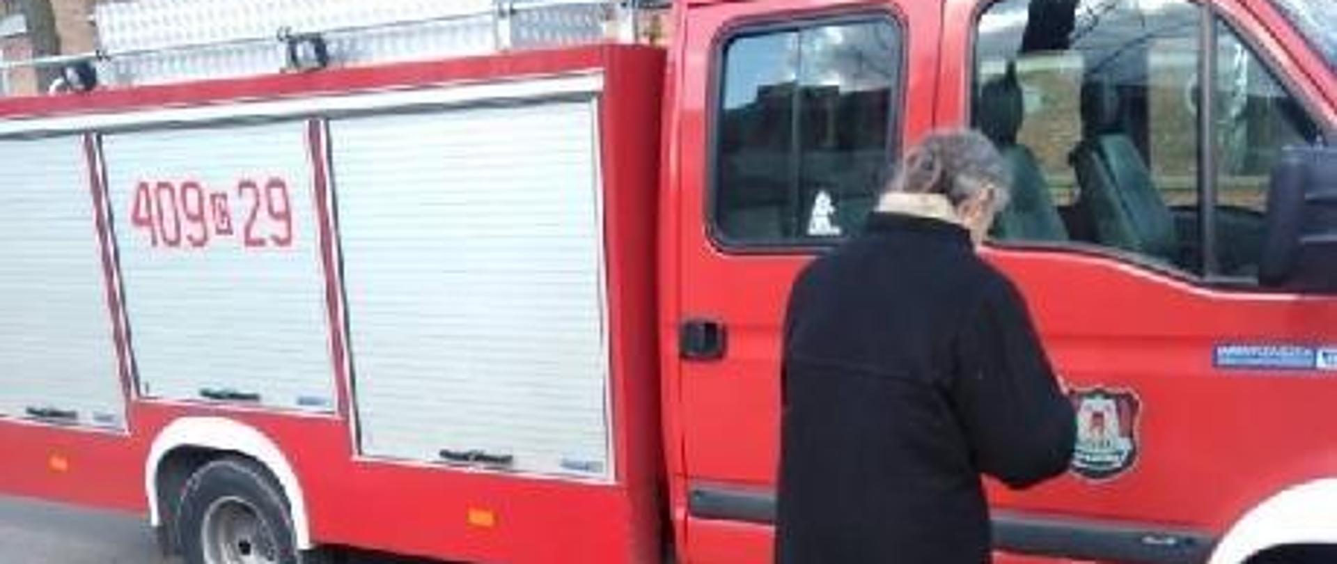 Zdjęcie przedstawia kobietę w czarnym płaszczu stojącą tyłem, na trawniku, na tle samochodu ratowniczo-gaśniczego należącego do OSP w Raciążku z widocznym numerem operacyjnym 409c29. W tle widoczna część kościoła z czerwonej cegły.