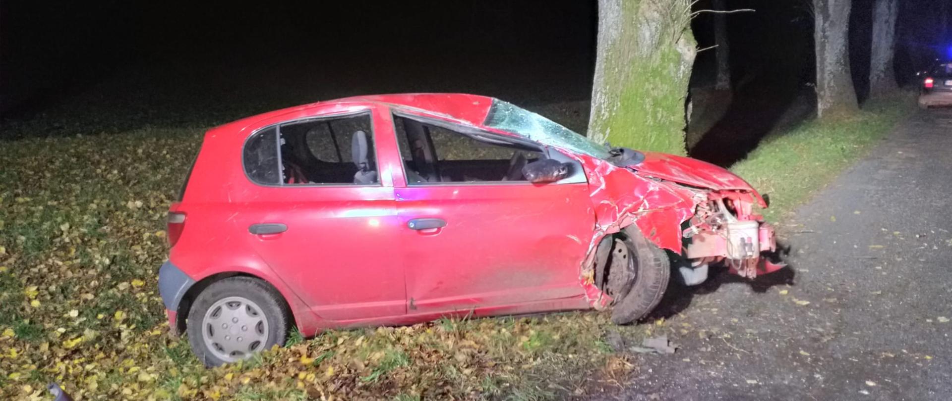 na zdjęciu czerwone auto osobowe uderzyło w przydrożne drzewo. Pojazd na poboczu drogi. 