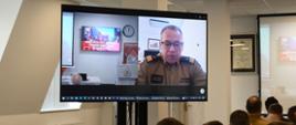 Na monitorze wyświetlane jest połączenie z Komendą Główną Państwowej Straży Pożarnej obok siedzą na krzesłach strażacy podczas narady. 