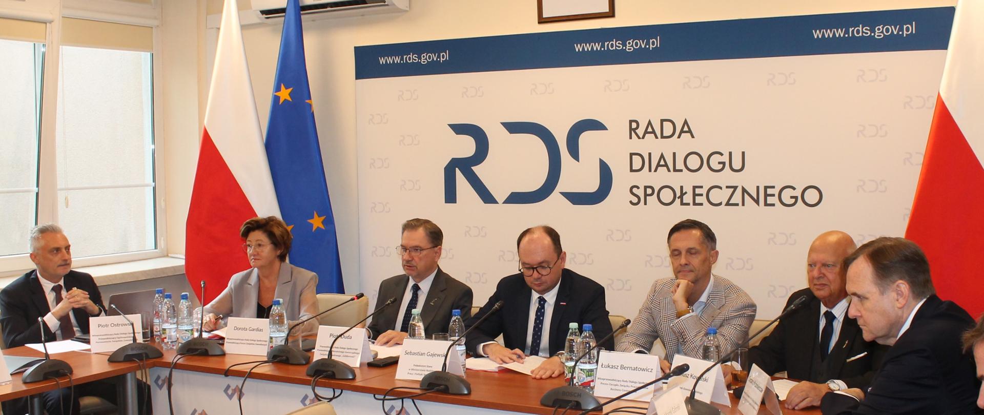 Na zdjęciu widnieje stół prezydialny za którym siedzą od lewej strony: Piotr Ostrowski – Przewodniczący Ogólnopolskiego porozumienia Związków Zawodowych, Dorota gardias – Przewodnicząca Forum Związków Zawodowych, Piotr Duda – przewodniczący Rady Dialogu Społecznego, Sebastian Gajewski – Podsekretarz Stanu w Ministerstwie Ministra Rodziny, Pracy i Polityki Społecznej, Łukasz Bernatowicz – Business Centre Club, Janusz Kowalski – Związek Rzemiosła Polskiego, Marek Górski – Prezydent Konfederacji Lewiatan. W tle siedzących dwie flagi: UE i Polski oraz baner z napisem Rada Dialogu Społecznego.