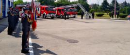 Fotografia przedstawia na pierwszym planie po lewej stronie poczet sztandarowy składający się z trzech strażaków, jeden z nich trzyma sztandar Komendy Miejskie Państwowej Straży Pożarnej w Sosnowcu. Za pocztem widać funkcjonariuszy państwowej straży pożarnej w umundurowaniu wyjściowym stojących w dwuszeregu podczas uroczystej zbiórki. Na dalszym planie widać pojazdy pożarnicze ustawione obok siebie. Po prawej stronie w oddali widać maszt z flagą rzeczypospolitej polskiej.