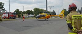 Wspólna akcja strażaków i LPR. Parking jednego z rawickich hipermarketów. Na nim śmigłowiec Lotniczego Pogotowia Ratunkowego, którego załoga przygotowuje się do startu. Z lewej strony wóz straży pożarnej. W tle drzewa, budynki i samochody.