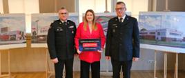 Jasne pomieszczenie, w którym na tle zdjęć z wizualizacją budynku stoi dwóch strażaków w mundurach galowych a pomiędzy nimi kobieta w różowej marynarce.
