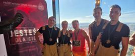 Grupa strażaków i uczestnik zawodów sportowych pozuje do wspólnego zdjęcia przy oknie. Strażacy mają na sobie spodnie ochronne i koszulki, a jedna z osób trzyma medal zawieszony na szyi. W tle widoczna jest panorama miasta oraz baner promocyjny Państwowej Straży Pożarnej.
