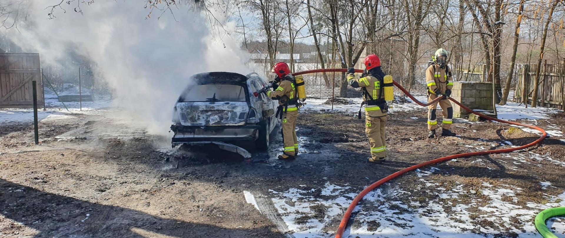 Na zdjęciu widać trzech strażaków w ubraniach specjalnych z założonymi na plecach aparatami ochrony dróg oddechowym. Strażacy gaszą palący się samochód osobowy na prywatnej posesji. W rękach trzymają wąż gaśniczy. Z samochodu wydobywa się gęsty, biały dym. Na ziemi zalega nieznaczna warstwa śniegu.