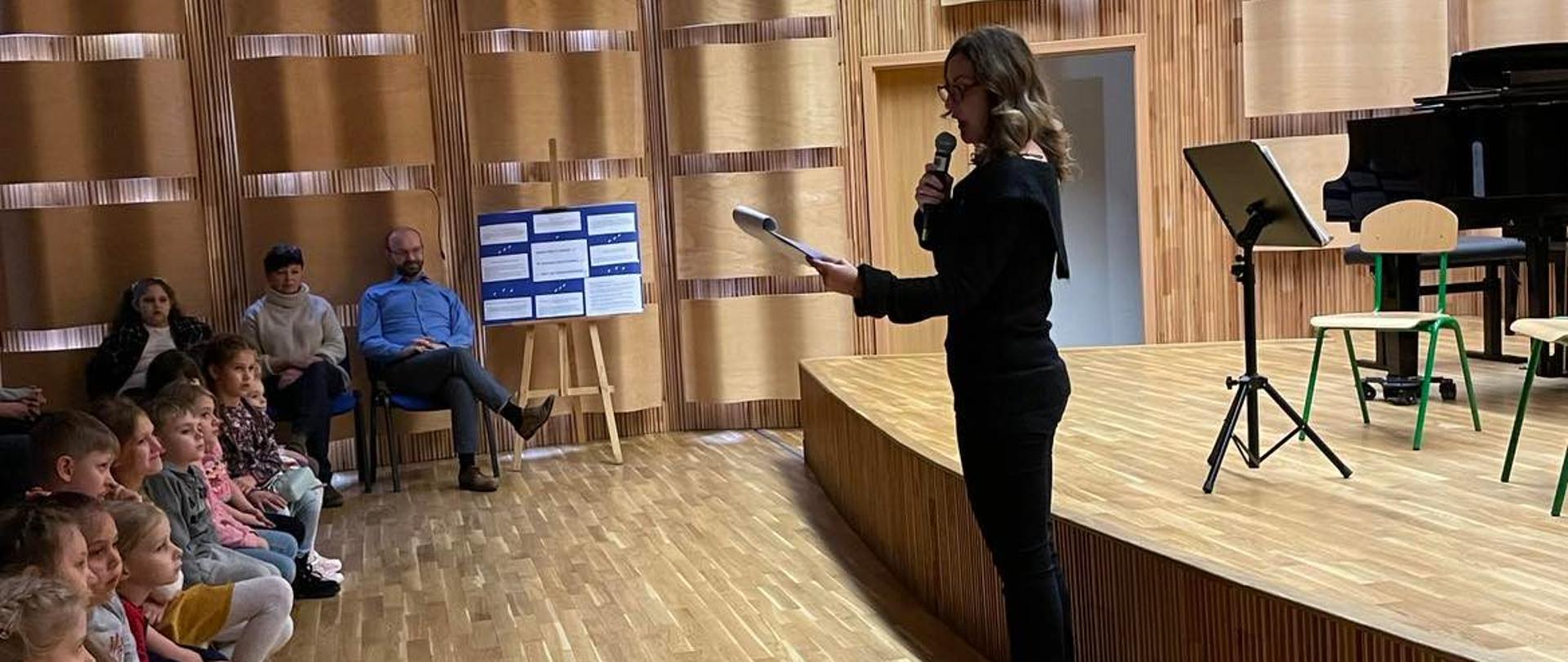 Zdjęcie sali koncertowej. Po lewej stronie siedzą dzieci na ławeczce, na środku dyrekcja siedzi na krzesłach, po prawej stoi nauczycielka i zapowiada utwory w czasie koncertu.