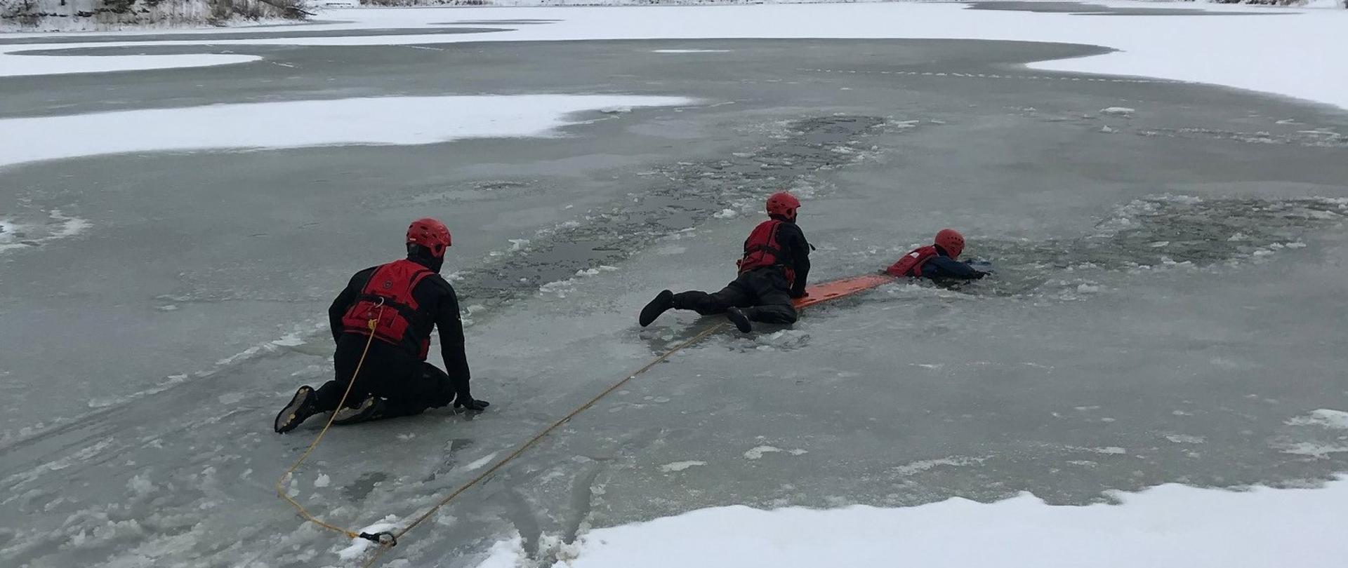 Jezioro pokryte lodem. W przeręblu - w wodzie, pływa ratownik ubrany w skafander oraz kask ochronny. Obok niego na tafli lodowej znajduje się ratownik, który próbuje go wyciągnąć z wody podając mu deskę. Deska jest zabezpieczona linką, do której przypięty jest również drugi ratownik zabezpieczający działania. 