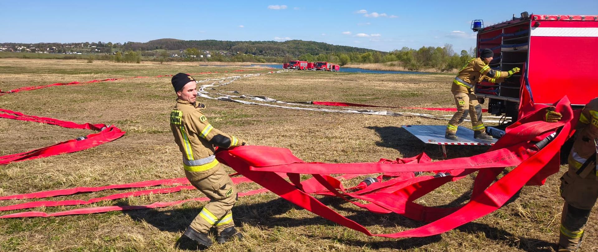 Strażak, który ciągnie węże pożarnicze. Strażak w ubraniu specjalnym, wąż czerwony, bez wody, suchy. Za nim fragment pojazdu wężowego, a z tyłu, na jego platformie stoi drugi strażak. W tle daleko widać pojazdy pożarnicze, wzniesienia i las.