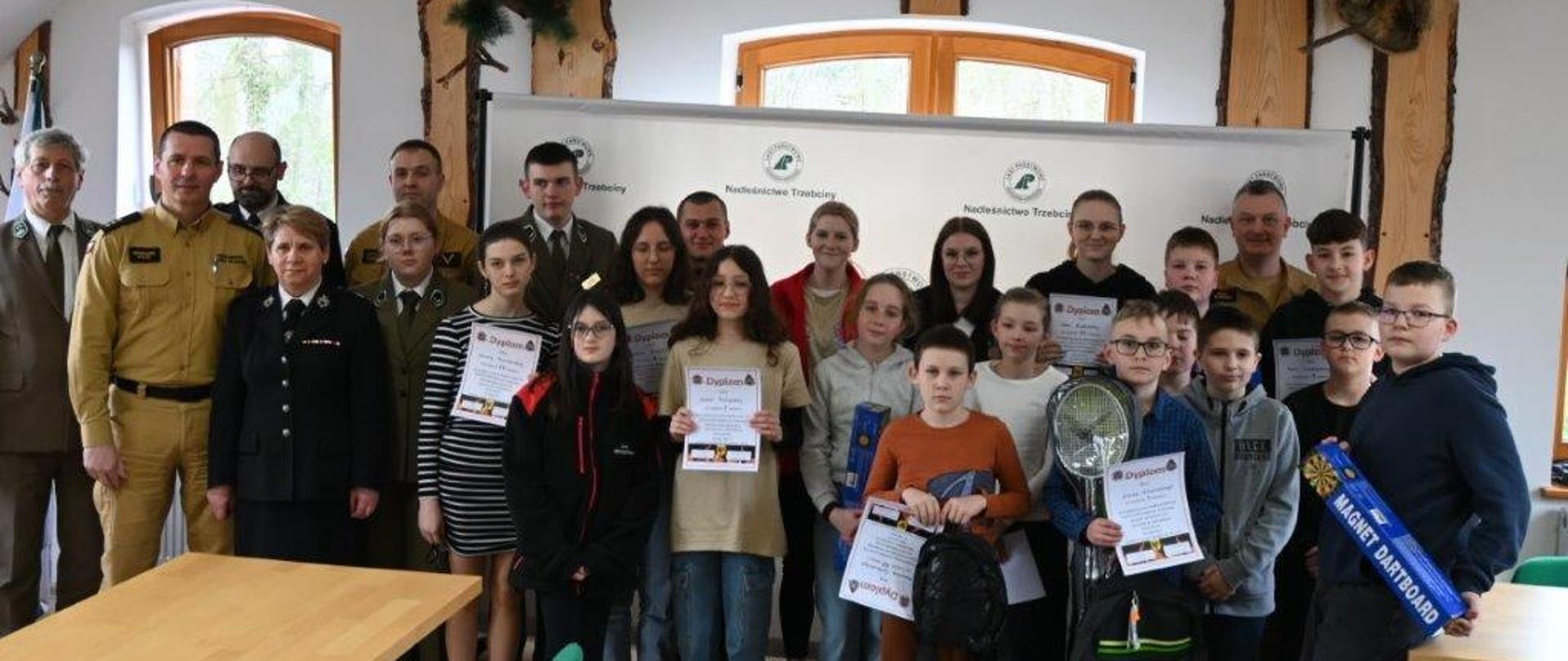 Grupa osób stoi. Z przodu młodzież trzyma dyplomy. Za dziećmi stoją osoby dorosłe. Wśród osób dorosłych stoją funkcjonariusze PSP w żółtych mundurach. Za grupą osób stoi biała tablica. Za tablicą na ścianie wisi godło Polski, sztuczne ptaki oraz krzyż. Na ścianie za ludźmi są widoczne okna. Ściany są białe z elementami drewna. Przed grupą ludzi po lewej stronie widać stolik i krzesło. 