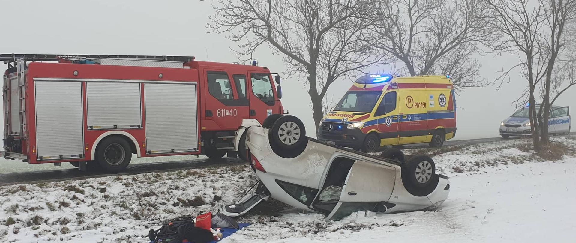 Samochód osobowy stojący na dachu na poboczu drogi. W tle samochody służb: straż pożarna, policja, pogotowie ratunkowe