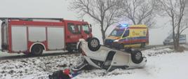Samochód osobowy stojący na dachu na poboczu drogi. W tle samochody służb: straż pożarna, policja, pogotowie ratunkowe