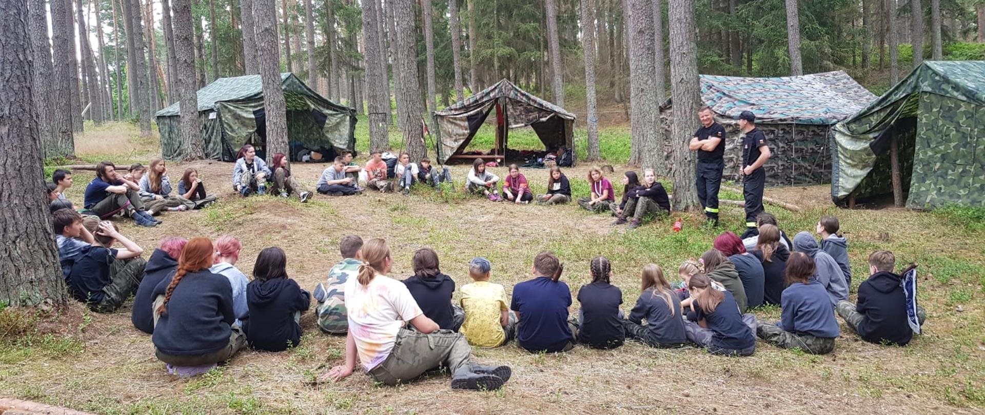 Na zdjęciu widać grupę młodzieży siedzącej na ziemi w kółku. Po prawej stronie widać dwóch strażaków prowadzących prelekcję nt. bezpiecznego zachowania się w terenie leśnym i nad wodą. W tle widać namioty i drzewa.