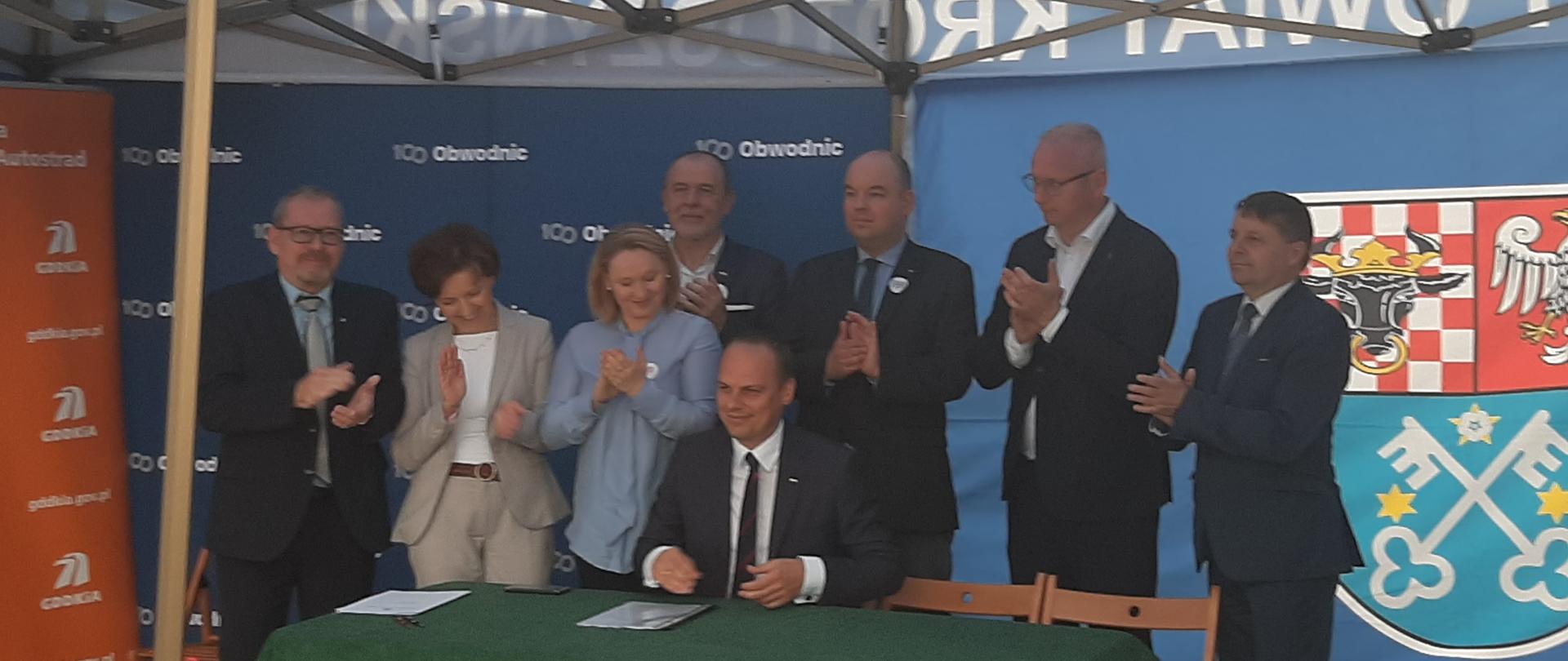 Na zdjęciu widać grupę ludzi, która stoi przy osobie podpisującej dokumenty. Widać ruchy uczestników, którzy klaszczą brawo okazując tym zadowolenie.