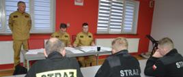 Egzamin KDR. Świetlica w Komendzie Powiatowej PSP w Rawiczu. Na przeciwko siebie, za stołami siedzą członkowie komisji egzaminacyjnej oraz trzej egzaminowani druhowie w czarnych ubraniach koszarowych. Na stole komisji znajduje się dokumentacja szkoleniowa. W tle okna.