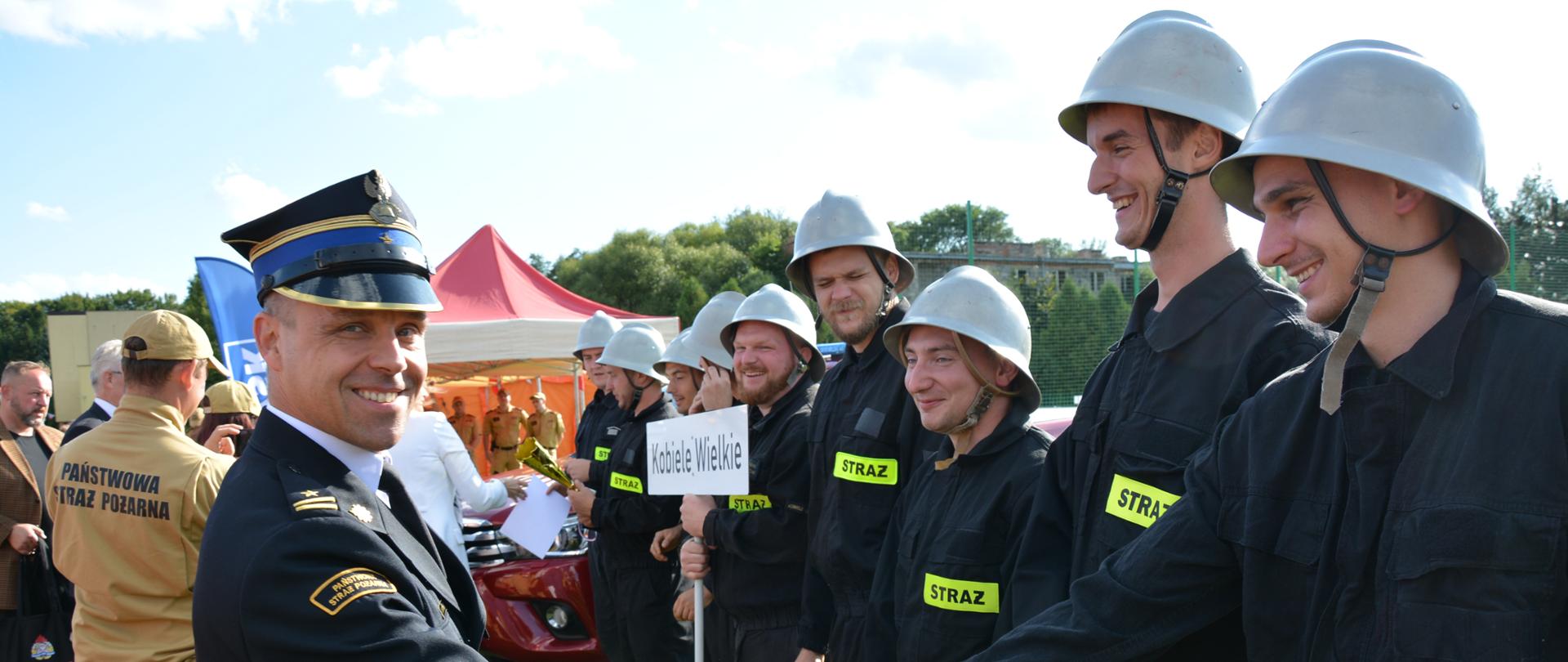 unkcjonariusz PSP w mundurze galowym podaje rękę jednemu z zawodników ustawionej w szeregu drużyny. Strażacy ubrani są w czarne mundury i charakterystyczne srebrne hełmy. W środku grupy widoczna jest tablica „Kobiele Wielkie”. Zdjęcie pokazuje bezpośredni moment składania gratulacji uczestnikom.