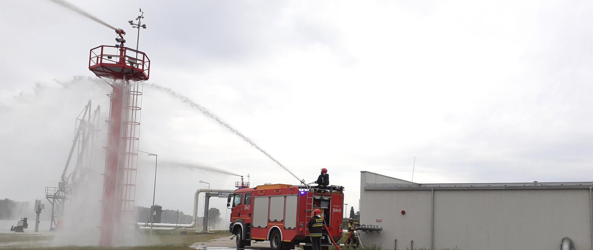 Na zdjęciu widać wóz bojowy straży pożarnej oraz dwóch strażaków z których jeden obsługuje działko na samochodzie a drugi obsługuje autopompę oraz monitor gaśniczy. Podawane prądy gaśnicze piany ciężkiej. Z prawej strony budynek zasilający i sterujący infrastrukturą gaśniczą nabrzeża 