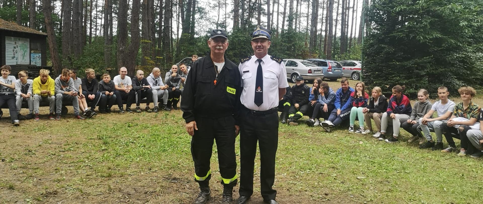 Z prawej Komendant Miejski PSP w Bydgoszczy st. bryg. Janisław Buller w białej koszuli munduru wyjściowego, z lewej dh Leszek Szmyt prezes Zarządu Oddziału Miejskiego ZOSP RP w Bydgoszczy w czarnym ubraniu koszarowym, w tle na ławkach uczestnicy obozu.