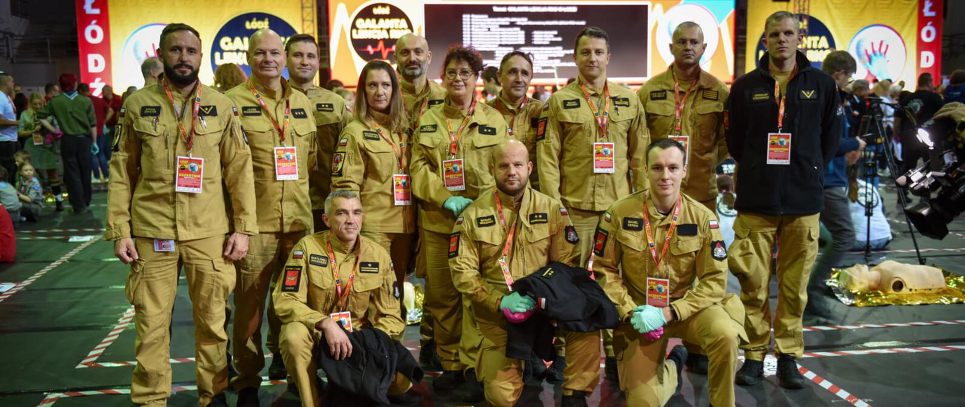 Grupa ratowników w beżowych mundurach pozuje do zdjęcia na tle sceny, przed nimi na ziemi leży manekin niemowlęcia na złotej folii termicznej.