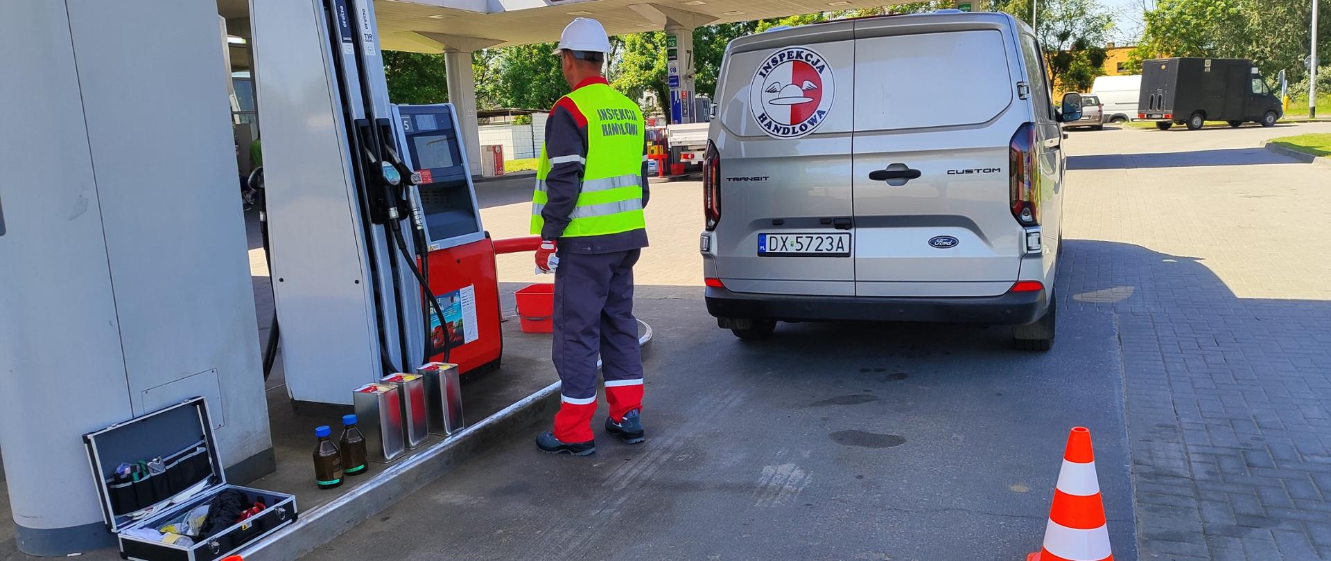 Pracownik Inspekcji Handlowej w trakcie czynności kontrolnych na stacji paliw stoi przed dystrybutorem. Pod dystrybutorem stoją 3 metalowe pojemniki na próby i 2 szklane butelki na próby oraz walizka ze sprzętem. Obszar pracy jest odgrodzony pachołkami, w tle samochód Inspekcji Handlowej do przewożenia paliw i infrastruktura stacji. 