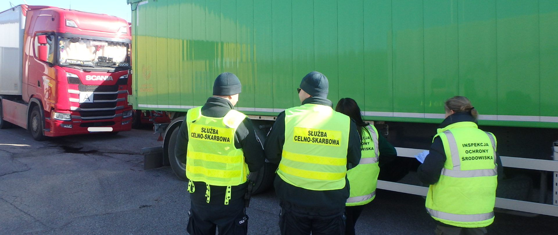 Widoczne są cztery osoby obrócone plecami stojące na ulicy (dwóch mężczyzn ubranych w czapki i kamizelki z napisem "Służba Celno-Skarbowa" oraz dwie kobiety z kamizelkami z napisem "Inspekcja Ochrony Środowiska") ustawieni przed zaparkowanymi samochodami ciężarowymi.