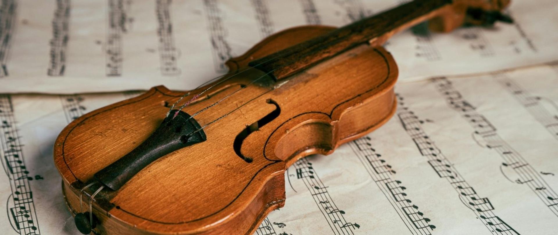 Plakat w pionie informujący o koncercie uczniów klasy skrzypiec p. Joanny Walickiej , który odbędzie się 12 marca o 17.30. Plakat w kolorystyce stonowanej, beżowej. Głównym elementem plakatu, umieszczonym pośrodku jest zdjęcie skrzypiec położonych na nutach. U góry i dołu plakatu białymi literami nazwa wydarzenia, data i godzina. 