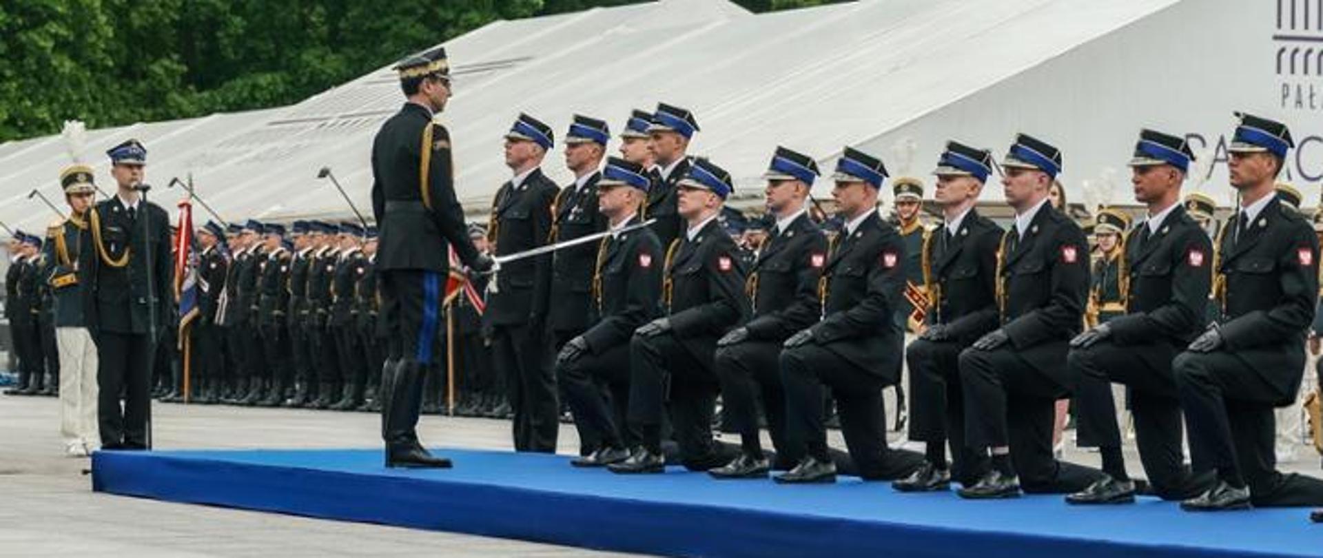 Zdjęcie przedstawia moment promocji. awansowani klęczą w rzędzie w mundurach galowych. generał straży szablą awansuje uderzając awansowanych w ramię. W tle białe namioty.