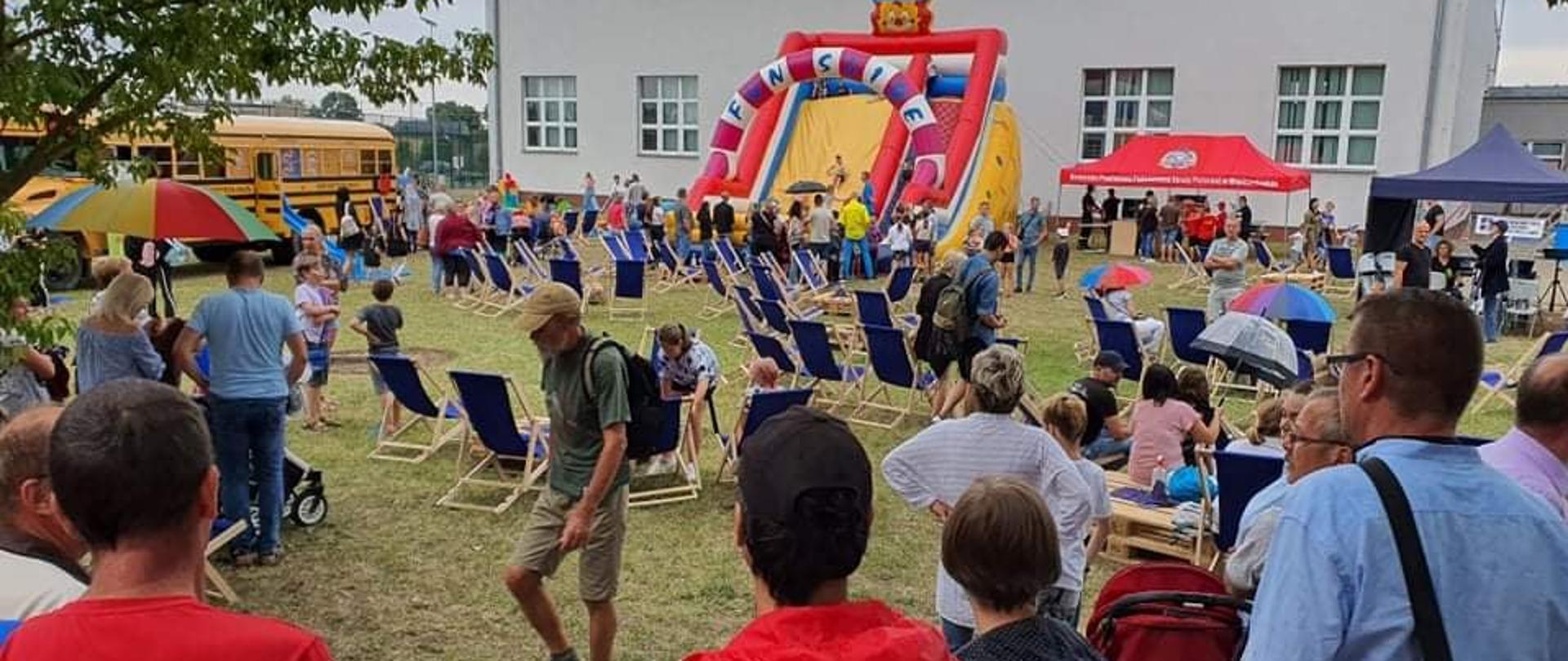 Zdjęcie przedstawia plac, gdzie odbywa się festyn na zakończenie lata. Dużo ludzi, rozłożone leżaki, w oddali dmuchaniec-zjeżdżalnia.