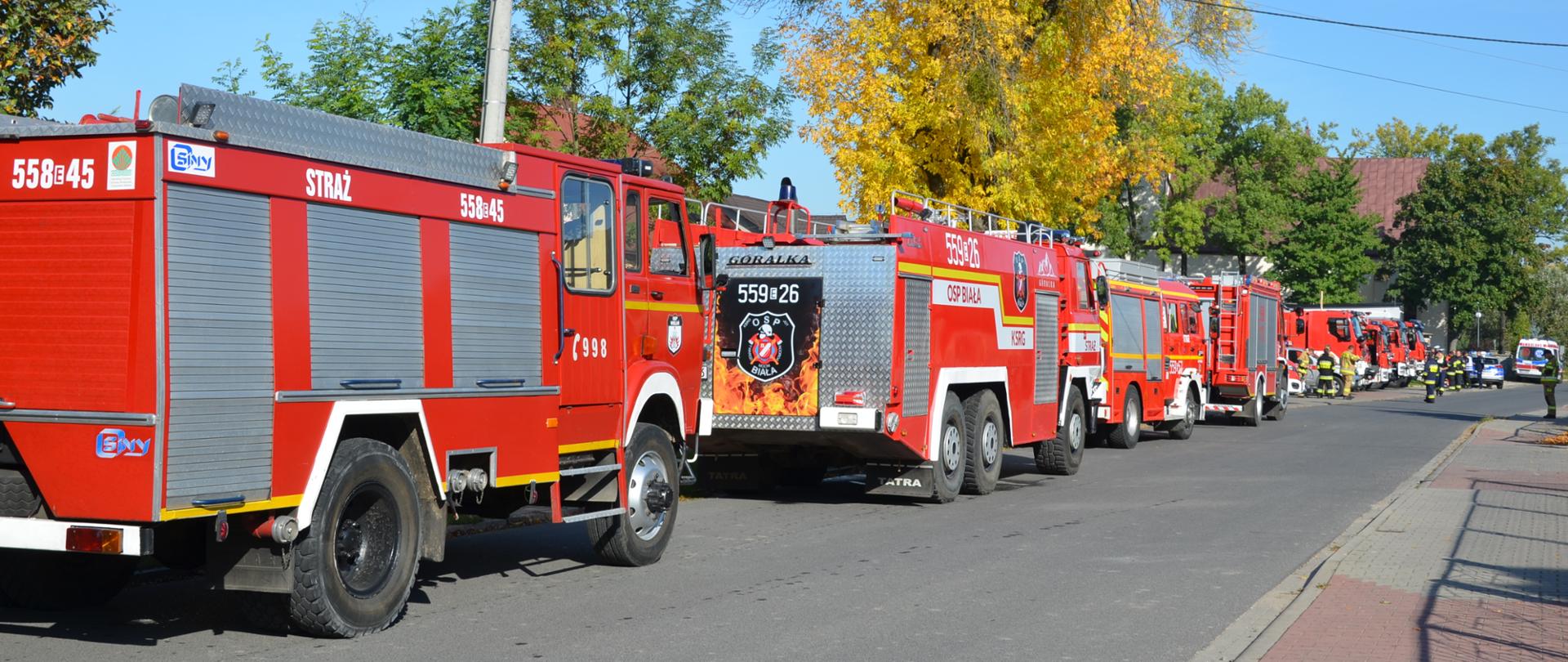 Zdjęcie przedstawia osiem samochodów pożarniczych koloru czerwonego ustawionych przy drodze, w tle karetka pogotowia oraz samochód policyjny, widoczni strażacy