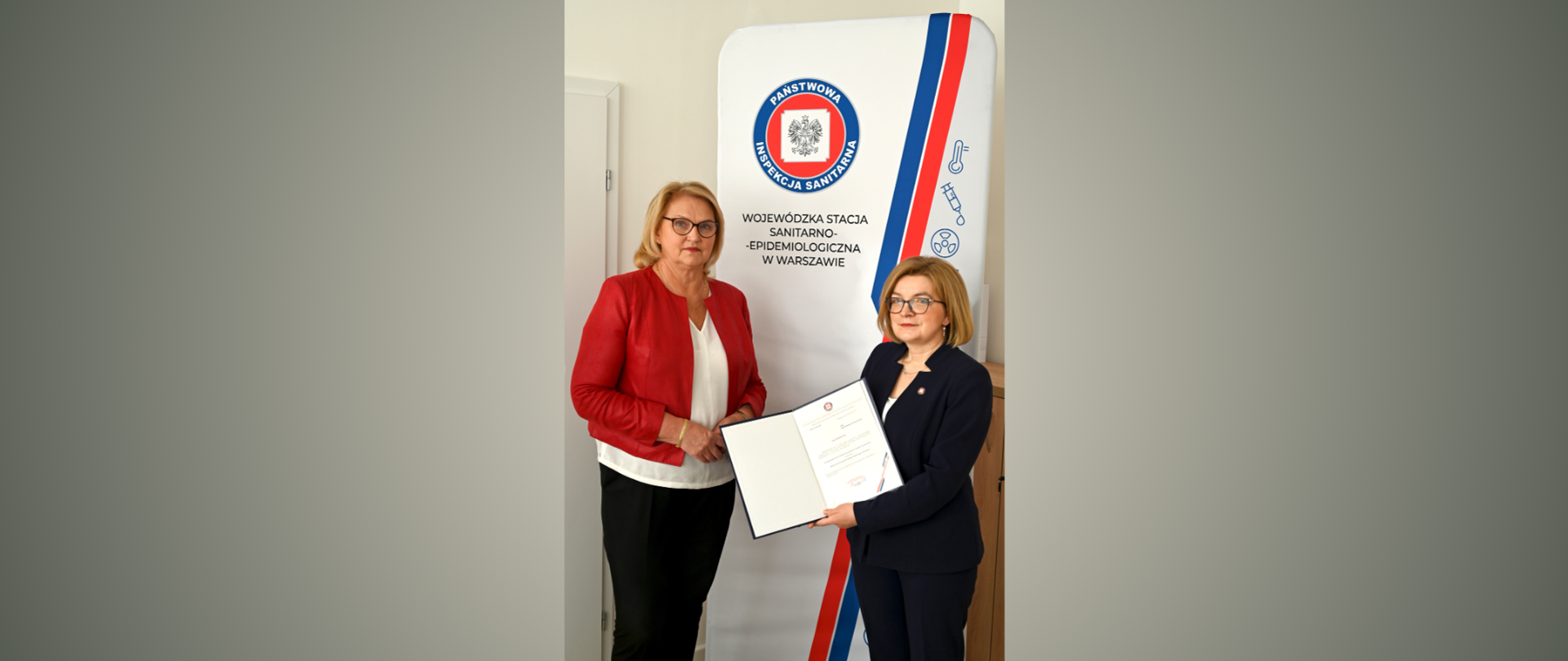 Dwie panie stojące obok siebie na tle logo Państwowej Inspekcji Sanitarnej. Jedna z nich trzyma akt powołania.